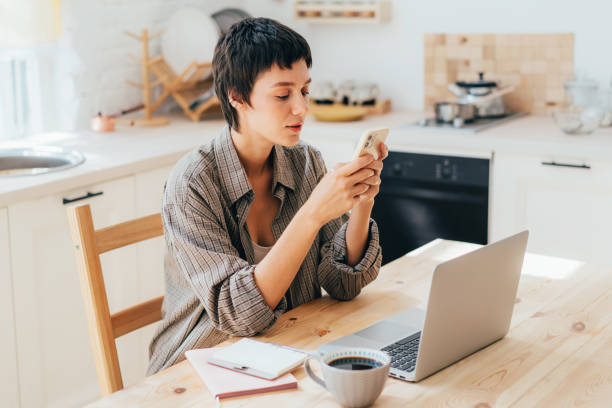 istock Young caucasian modern female freelancer sitting in the kitchen uses mobile phone and computer. 1424324253