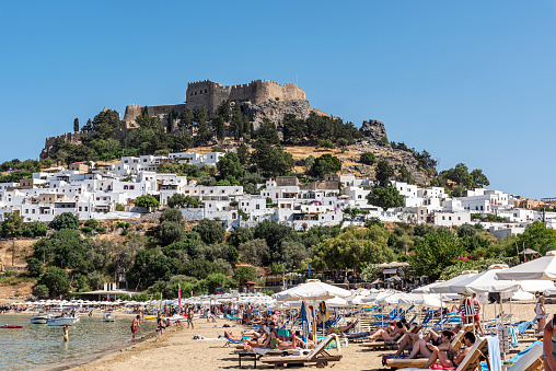 Rhodes Island, Greece - May 26, 2022: On the Greek island of Rhodes; The beach with people sunbathing and swimming by the sea in the foreground, the white houses of the famous town of Lindos and the Acropolis castle in the background.