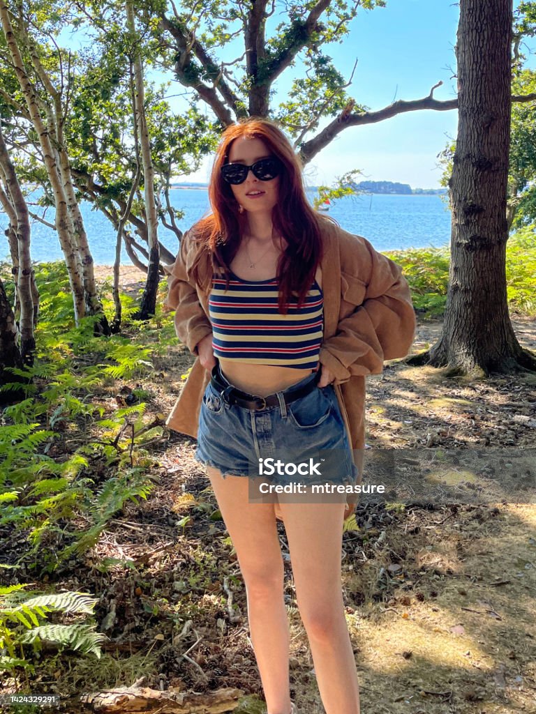 Image of woman with long, red hair pulling denim shorts with belt up by the waist, wearing jacket, striped vest top and mirrored sunglasses, standing in coastline woodland shade, looking at camera, blue summer sky, sea, focus on foreground Stock photo showing close-up view of long haired, redheaded woman standing in coastline woods beside the sea. 18-19 Years Stock Photo