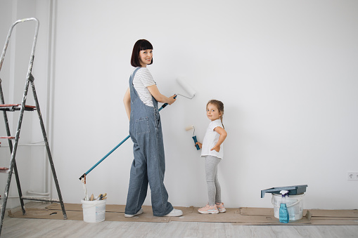 Full length portrait back view of young mother and her small cute daughter enjoying time together while painting wall.
