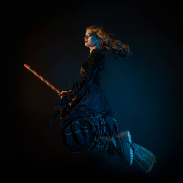A beautiful witch on a broomstick is flying to the sabbath. Young brunette woman in a dress and with a broom, rushing to a Halloween party, fire lighting, black background
