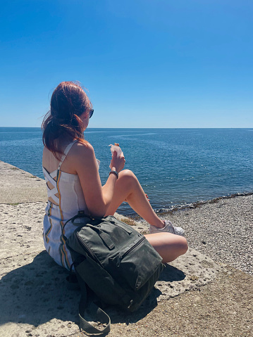 Stock photo showing close-up view of long haired, redheaded woman sitting looking at sea and eating an ice cream cone on the Cobb in harbour at the seaside town of Lyme Regis, Dorset, England, UK.