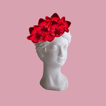 Female antique statue's bust with red lily flowers on her head isolated on a pink color background. Trendy collage in magazine surreal style. 3d contemporary art. Modern design