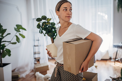 One woman, young woman carrying cardboard box and houseplant in her new home.