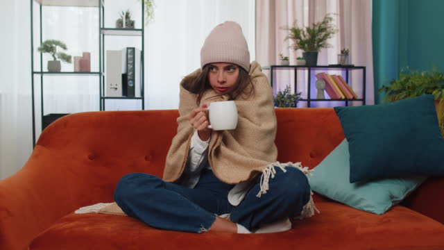 Sick young woman wear hat wrapped in plaid sit alone shivering from cold on sofa drinking hot tea