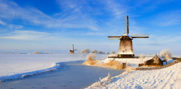 Snow covered windmills along a frozen canal in winter setting and a bit of ground fog. The location is Schermerhorn, Netherlands