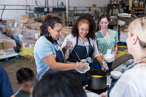 The diverse group of volunteers stand in an assembly line to serve soup bowls.