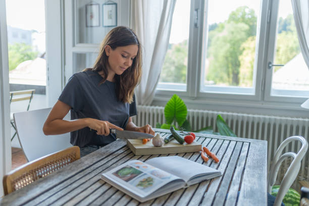 Young woman chops vegetables on kitchen table, recipe book