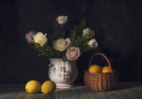Still life with roses in a vase and a yellow lemons in a wicker basket on a wooden table close-up