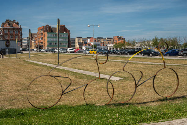 Europe, France, Dunkerque - July 9, 2022: Tour de France memorabilia. 2 rusted bicycle with rider profiles, 1 yellow shirt, on lawn of Place du Mynck under blue sky. Housing and cars in back