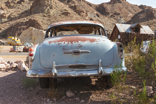 Nelson's Landing in Searchlight, NV USA, July 21, 2022: Image depicts rear of rusted, pastel blue, classic American vintage car with shiny chrome bumper and broken, missing windows. Car is parked within desert ghost town. Vintage automobiles can be seen parked in the distance.