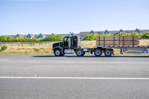 istock Black big rig semi truck tractor with extended cab transporting lumber cargo on flat bed semi trailer driving on the wide road along the apartments buildings 1424373020