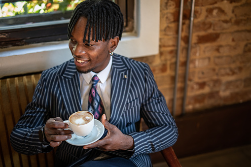 A young business man, of black race, elegantly dressed in a suit, sits in a cafe and drinks coffee, behind him is a brick wall.