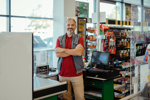 Portrait of mature adult male cashier looking at camera with crossed arms at supermarket