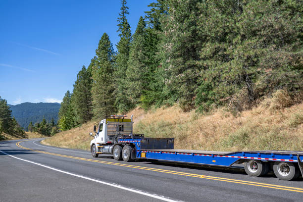 istock Day cab big rig semi truck tractor with empty low profile step down semi trailer for carry oversized loads running on the winding mountain road with forest on the hills 1424374019