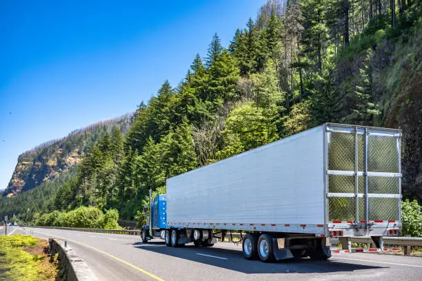 Photo of Classic blue American bonnet big rig semi truck transporting cargo in refrigerator semi trailer driving on the divided highway road with mountain and wild forest