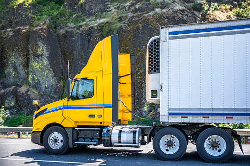 Day cab for local delivery bright yellow big rig semi truck with roof spoiler transporting food cargo in reefer semi trailer running on the mountain road with rock wall on the side in Columbia Gorge