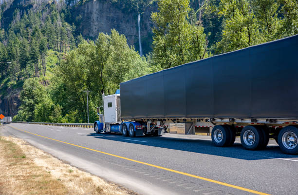 istock Extended cab white classic big rig semi truck tractor transporting cargo in tented black dry van semi trailer driving on the one way road with mountain and waterfall on the side in Columbia Gorge 1424376698