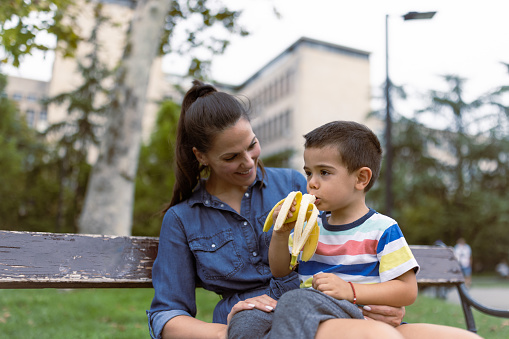 Mother feeding her little son. They are sitting in a park bench, eating banana