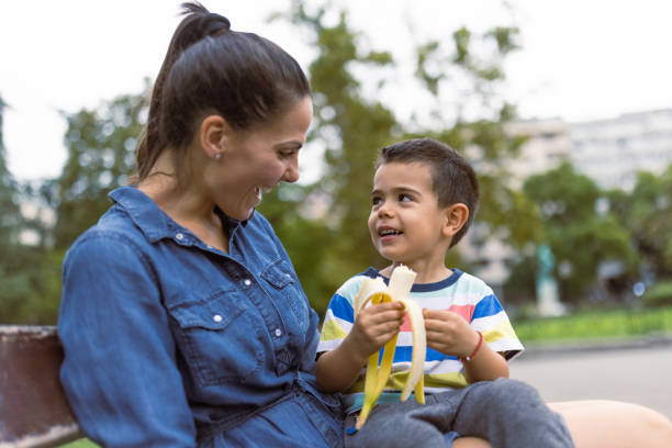 Mother feeding her little son. They are sitting in a park bench, eating banana