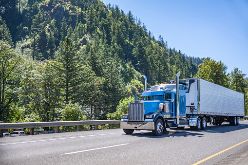 Classic blue long haul big rig semi truck tractor with chrome exhaust pipes transporting commercial cargo in refrigerator semi trailer moving on the highway road in Columbia Gorge National Reserve