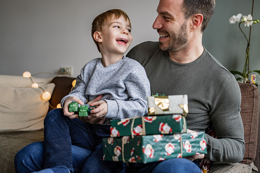 Cute boy sitting on his father's lap. They are holding Christmas presents and looking at each other.