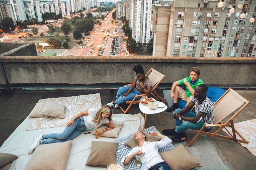 A multiracial group of tenants enjoys their time while watching a movie on the rooftop at dusk. In this photo, they are sitting or lying down.