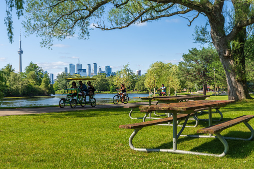 Toronto, Ontario, Canada - July 30 2021 : People cycling in the Toronto Islands Park Centre Island. Wooden Bench In Foreground, Toronto City Skyline in Background.