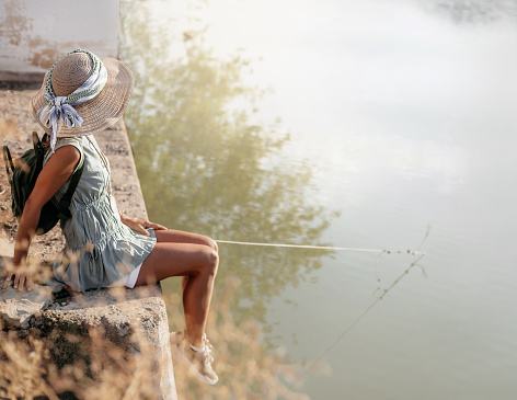 middle-aged woman in dress and hat sitting comfortably on the edge of a lake jetty at sunset. concept tranquility and relaxation. landscape format