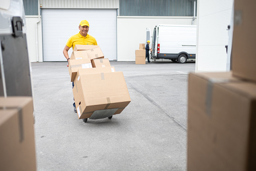 Warehouse worker moving hand truck loaded with cardboard boxes stack