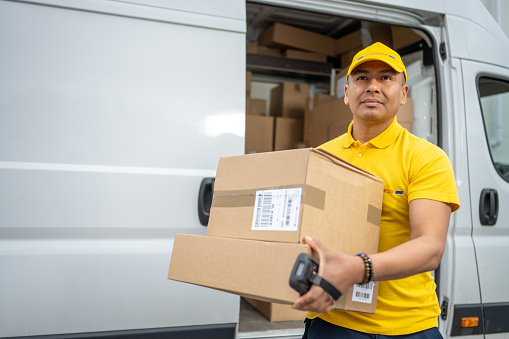 Delivery man in professional uniform carrying cardboard boxes stack, looking up. Courier unloading truck, holding order package and barcode reader