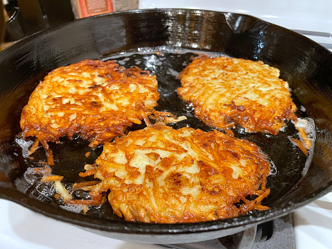 Black cast iron skillet with three golden brown potato latkes frying in oil for Hanukkah