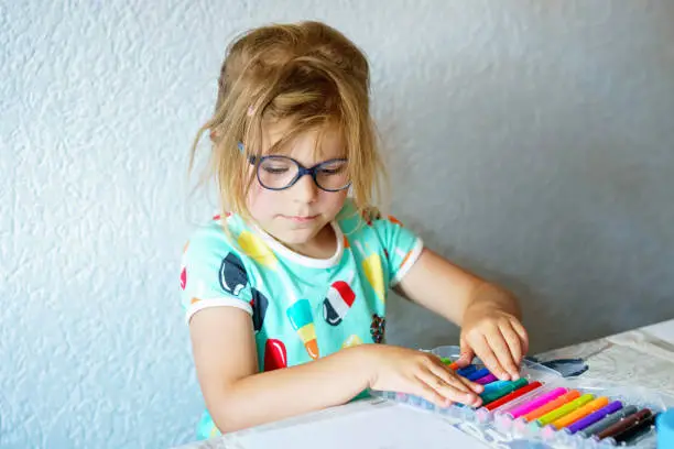 Happy preschool girl with glasses learning painting with colorful pencils and felt pens. Little toddler drawing at home on sunny summer day, using colorful feltpens. Creative activity for children