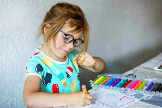 Happy preschool girl with glasses learning painting with colorful pencils and felt pens. Little toddler drawing at home on sunny summer day, using colorful feltpens. Creative activity for children