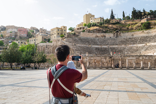 Man taking a photo of ancient Roman theater exterior in Amman downtown while carrying a baby boy in baby carrier on a sunny day in Jordan. Father and infant son traveling abroad