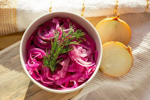 Delicious homemade marinated pickled purple onions in the white bowl with yellow onion bulbs in the kitchen.