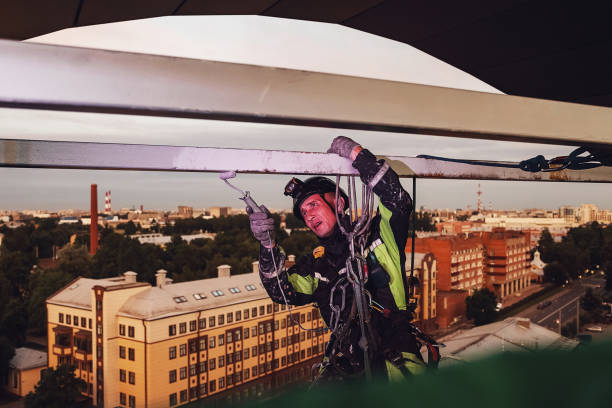 istock Industrial mountaineering worker in uniform hangs over building and paint roller construction at night urban background, painting facade. Rope access laborer on house. Industry dusk city work concept 1424503197