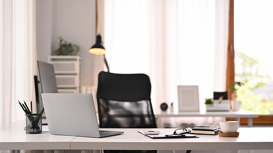 Horizontal image of laptop computer, documents and various office supplies on white table in modern interior.