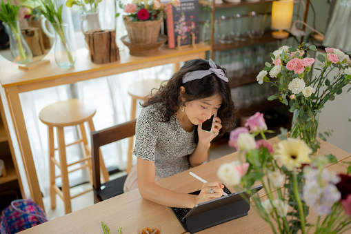 Young woman florist preparing for a new busy day using digital tablet and making business calls while taking customer orders.