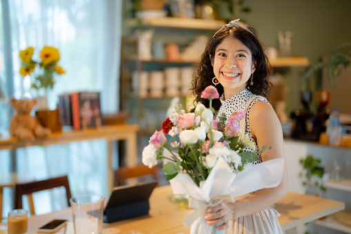Gorgeous florist woman wearing an apron arranging a bouquet of beautiful colorful flowers inside her floral shop while at the wooden counter.