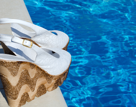 A pair of beautiful, snow-white, women's sandals against the backdrop of a blue pool. On a bright sunny day. Place for text.
