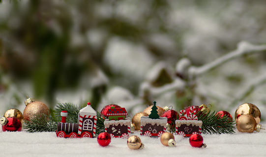A toy locomotive stands on the snow against the background of Christmas tree branches and Christmas balls, the background is blurred. Christmas, Christmas banner, postcard, flyer, layout design, narrow image