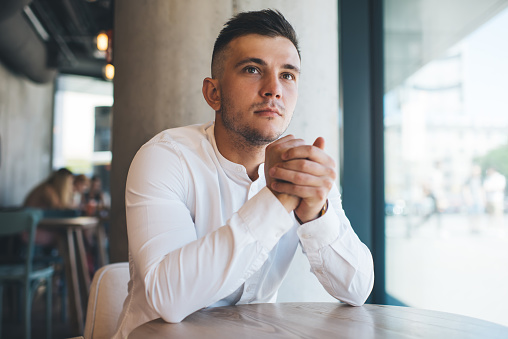 Young pensive male in white long sleeves tee looking away while sitting at empty wooden table in well lit stylish cafe in sunny day