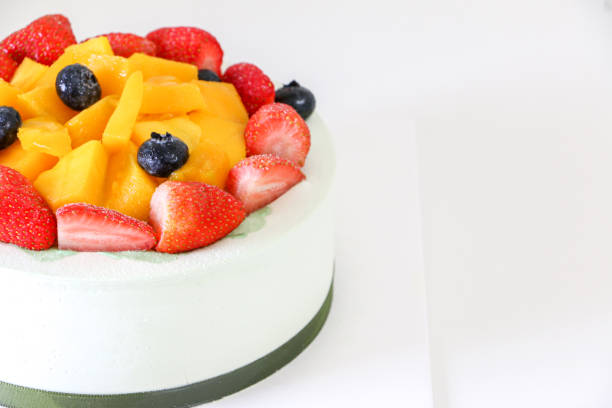 istock Creamy birthday cake, fruit cake, topped with fresh strawberries, blueberries and mango, on left of frame and white table 1424600757