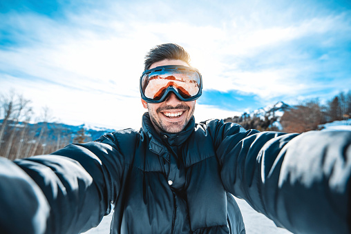 Young man taking selfie picture with smart cell phone hiking mountain on snowy slope - Happy skier having fun in ski resort vacation - Winter lifestyle, extreme sport and happy people concept