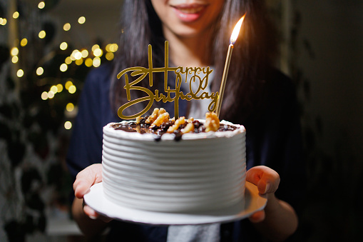 Cropped image of woman hands holding a beautiful birthday cake decorated with golden happy birthday sign and candle