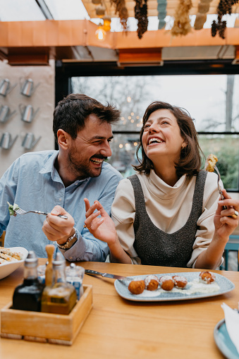 Spontaneous image of a beautiful brunet enjoying her lunch and a company of her handsome bearded partner, leaning at her, laughing. Both stylishly casually dressed while sitting at the restaurant