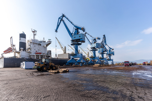 Shipping and Trucking Transportation Industry - tractor trailer sits in the foreground at a commercial dock. Containers are load on board a ship by cranes