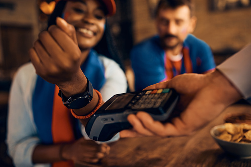 Close up of African American woman paying contactless with smart watch during sports championship in a bar.