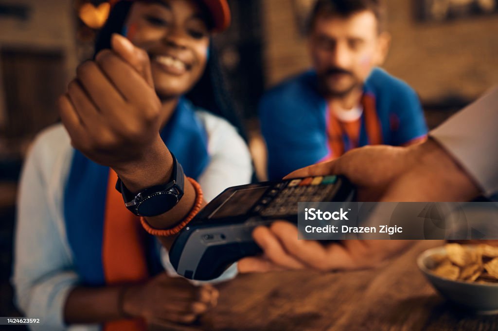 Close up of black sports fan using her smart watch while paying contactless in pub Close up of African American woman paying contactless with smart watch during sports championship in a bar. International Soccer Event Stock Photo
